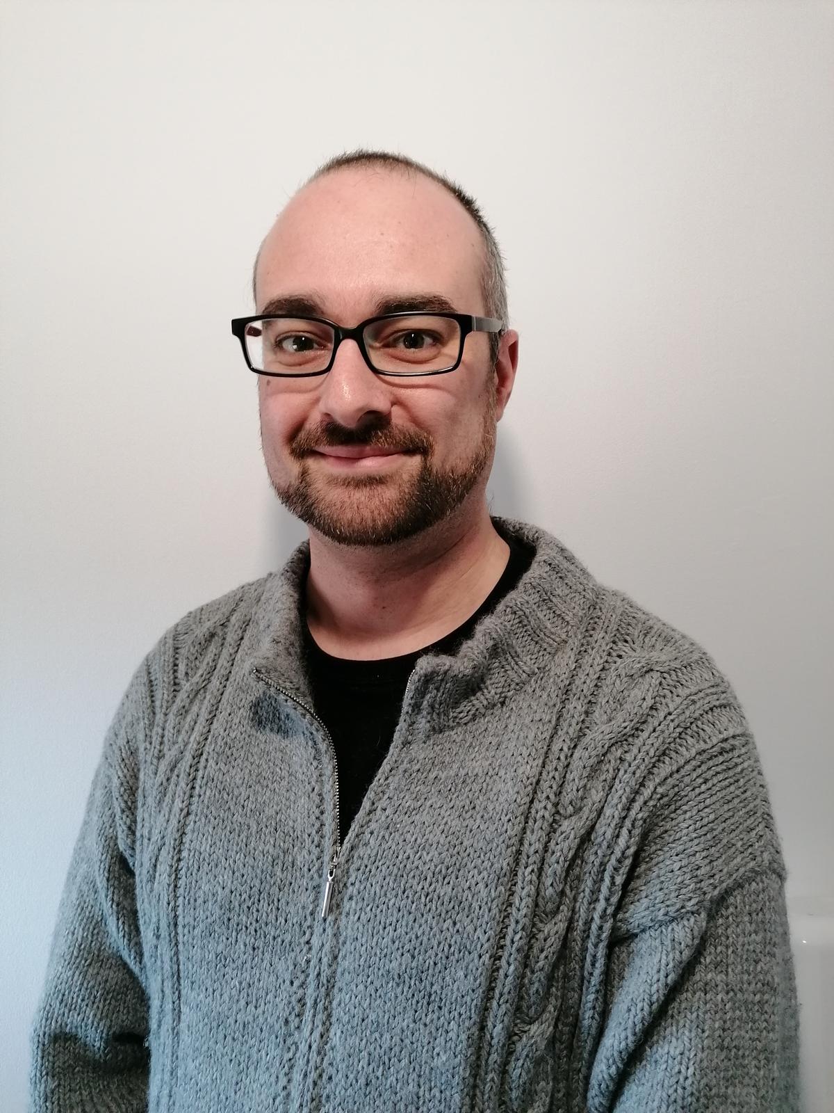 Picture of a (cute) caucasian white balding male with glasses, a trimmed beard, black t-shirt and a gray homemade sweater